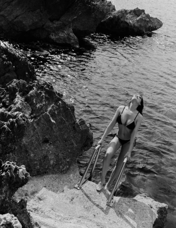 Black and white shot of a woman in a bikini climbing out of the ocean on a ladder, with rocks on the side, shot in natural light.