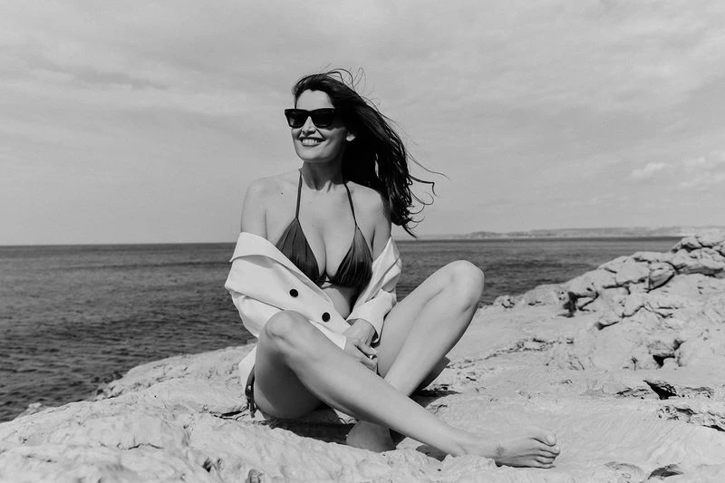 Woman in bikini and sunglasses smiles sitting on rocks near the ocean on a sunny day in a monochrome photograph.