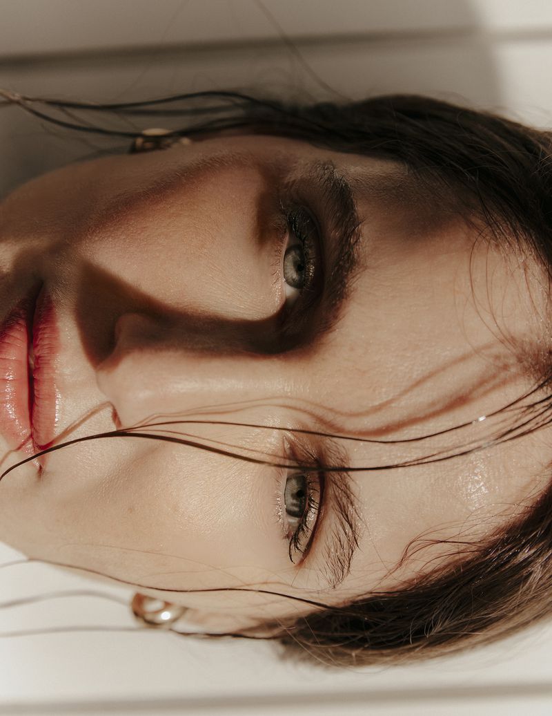 Close-up portrait of a woman with brown hair, light skin, and light-green eyes, featuring visible glossy lips and gold hoop earrings.