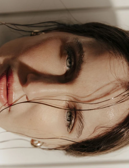 Close-up portrait of a woman with brown hair, light skin, and light-green eyes, featuring visible glossy lips and gold hoop earrings.