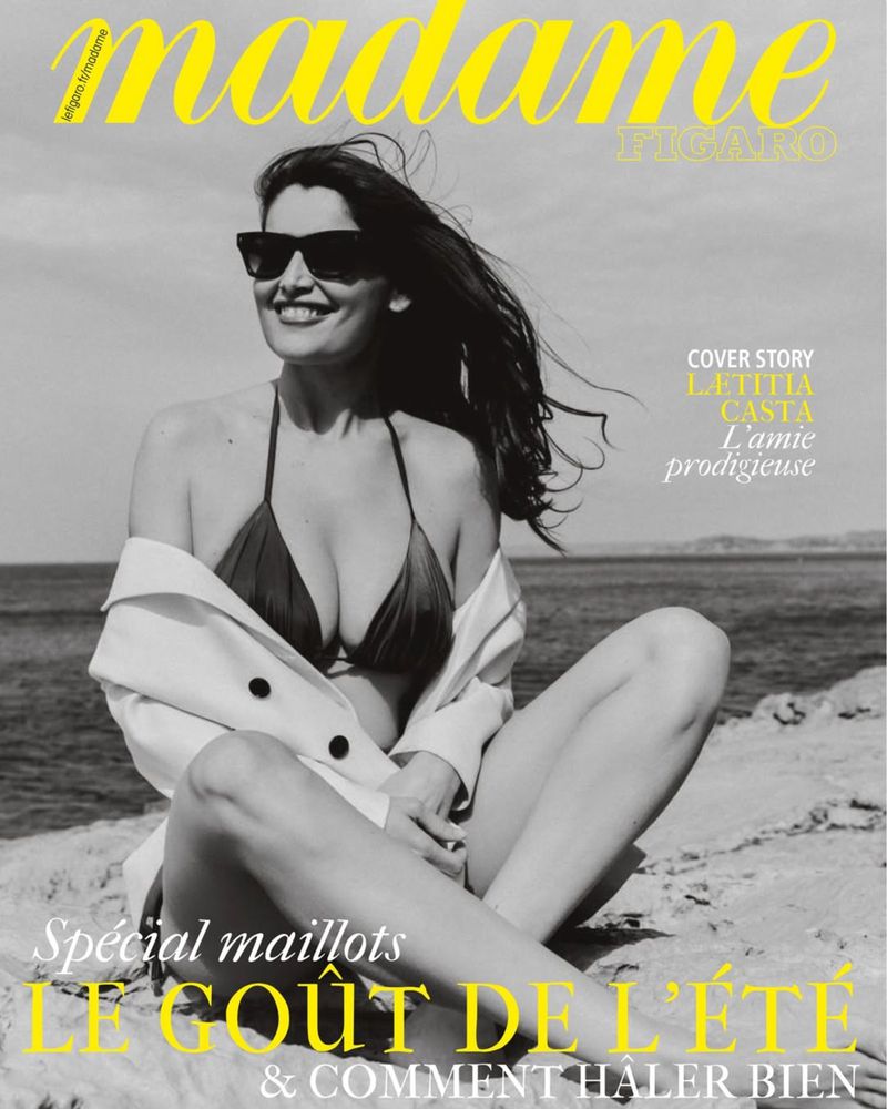 Letitia Casta on Madame Figaro magazine cover, wearing a bikini and jacket. Seated on rocks with ocean backdrop in a black and white photograph.