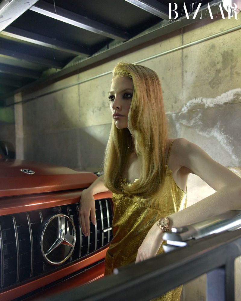 Fashion shot of a blonde woman in a metallic dress leaning on an orange SUV in a dimly lit garage, featuring BAZAAR magazine logo.