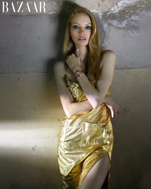 Fashion editorial shot of a fair-skinned adult woman in a gold dress with long blond hair, posing against a textured grey wall. 