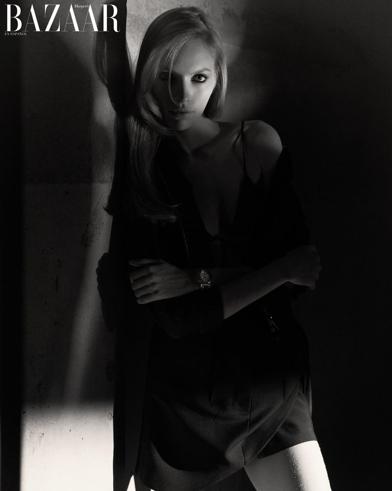 Blonde woman in dark outfit poses against a textured wall in a black and white fashion editorial, watch visible on wrist.