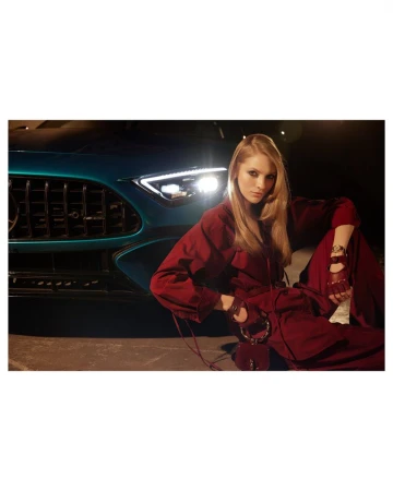 Blonde woman in red jumpsuit posing next to luxury car with teal finish in an indoor photo studio, eye-level angle.