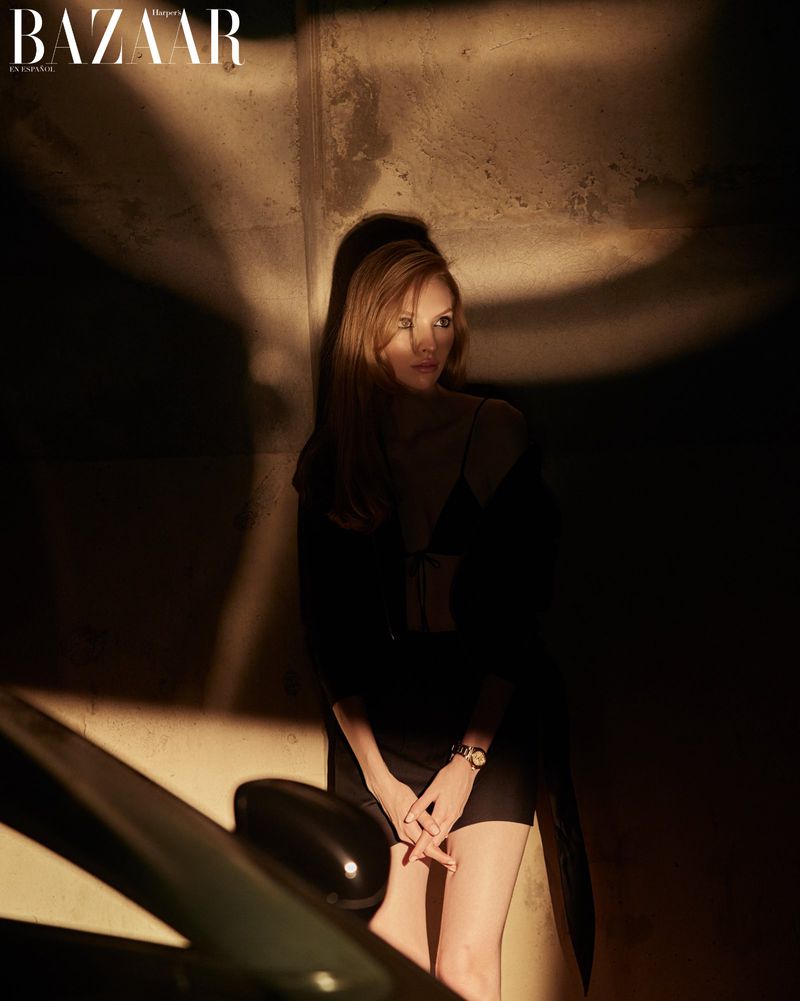 Woman poses near a car in a dramatic fashion editorial. The lighting is moody, emphasizing shadows on a concrete wall background.