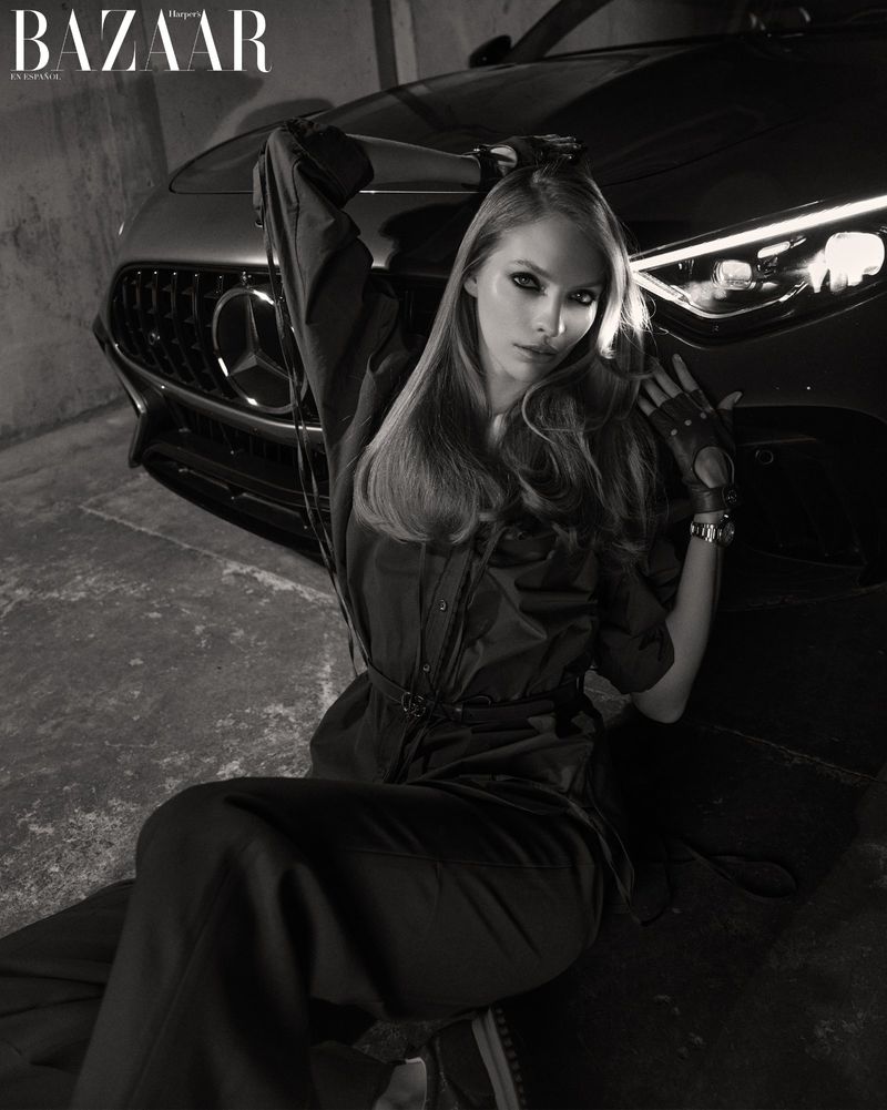 Monochrome fashion shot of a woman in a jumpsuit sitting in front of a luxury car with moody lighting and a concrete background.