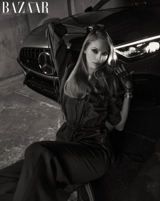 Monochrome fashion shot of a woman in a jumpsuit sitting in front of a luxury car with moody lighting and a concrete background.