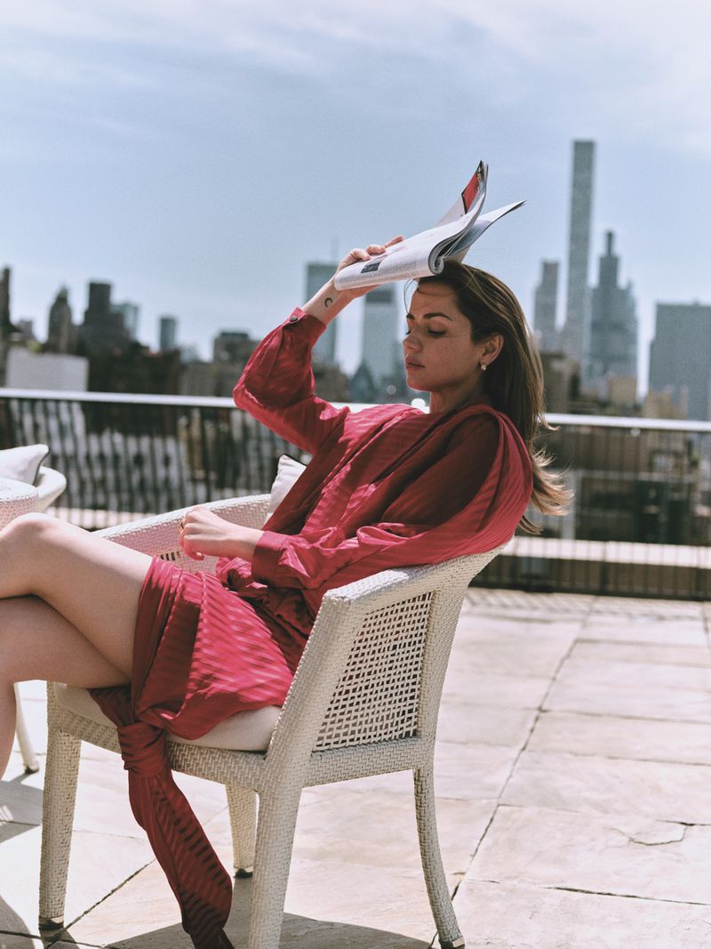 Ana de Armas seated in a white wicker armchair on a sunlit rooftop terrace, holding a magazine over her head against a city skyline.