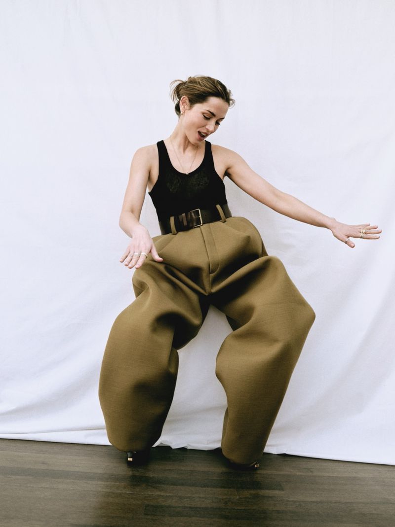 A woman poses indoors wearing oversized, wide-legged olive-green pants, a black tank top, and a dark belt against a white backdrop.