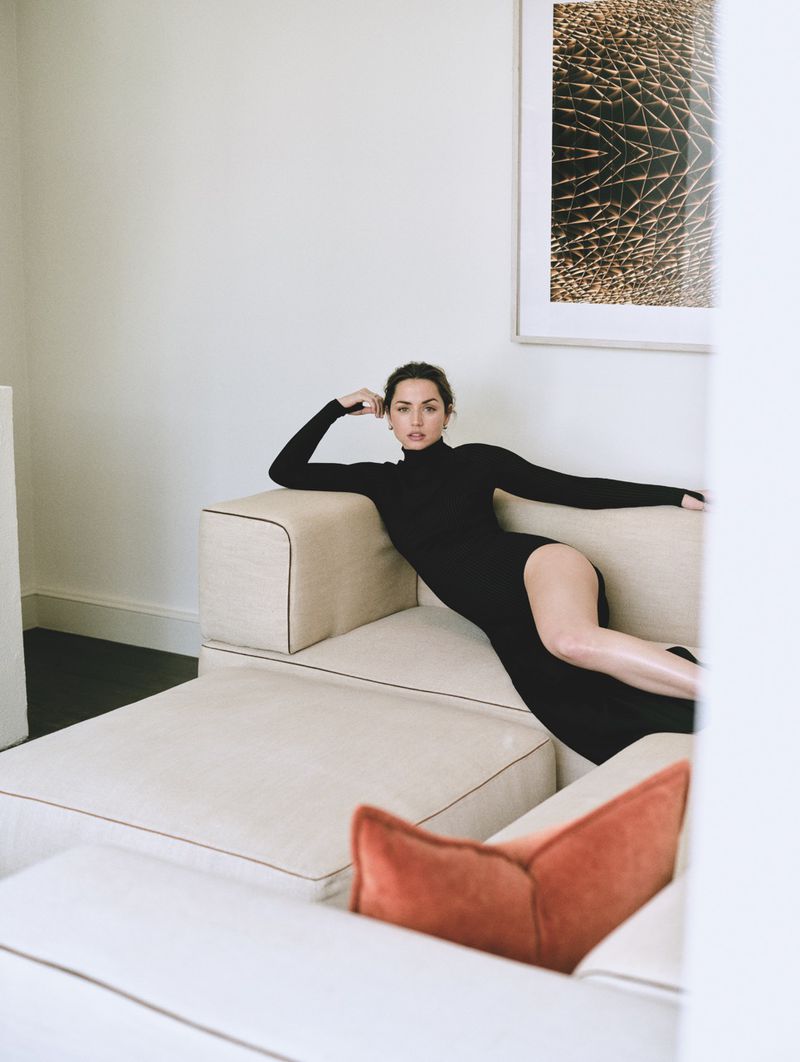 Ana de Armas poses on a light beige sofa in a long-sleeved black dress indoors with a framed print hanging on the wall behind.