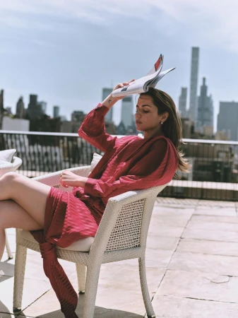 Ana de Armas seated in a white wicker armchair on a sunlit rooftop terrace, holding a magazine over her head against a city skyline.