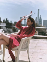 Ana de Armas seated in a white wicker armchair on a sunlit rooftop terrace, holding a magazine over her head against a city skyline.