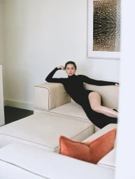 Ana de Armas reclines on a beige sectional sofa wearing a long-sleeve black turtleneck dress in a minimalist, bright interior.