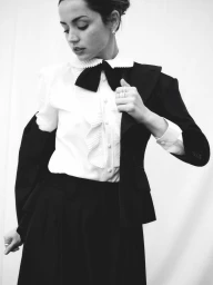 Ana de Armas wearing a ruffled white shirt with a black bow tie and black blazer against a light background.