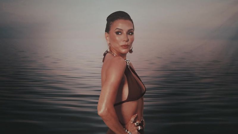 Adult female in brown bikini posing in front of a water background with dramatic light, wearing large hoop earrings and hand jewelry.