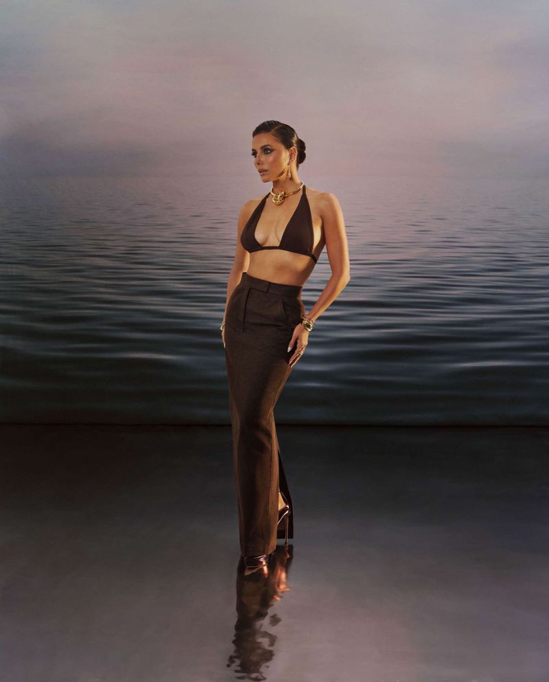 A woman in a brown two-piece outfit with gold jewelry stands against a blurred water backdrop in a full shot.