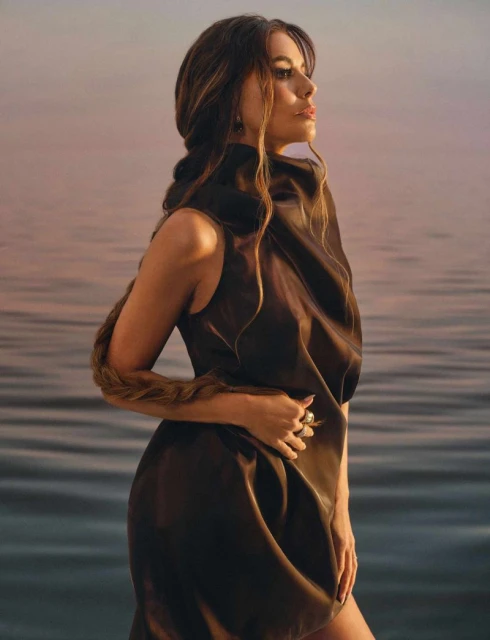 Woman in draped brown dress with long braid poses outdoors against water and sky at sunset for fashion or beauty content.