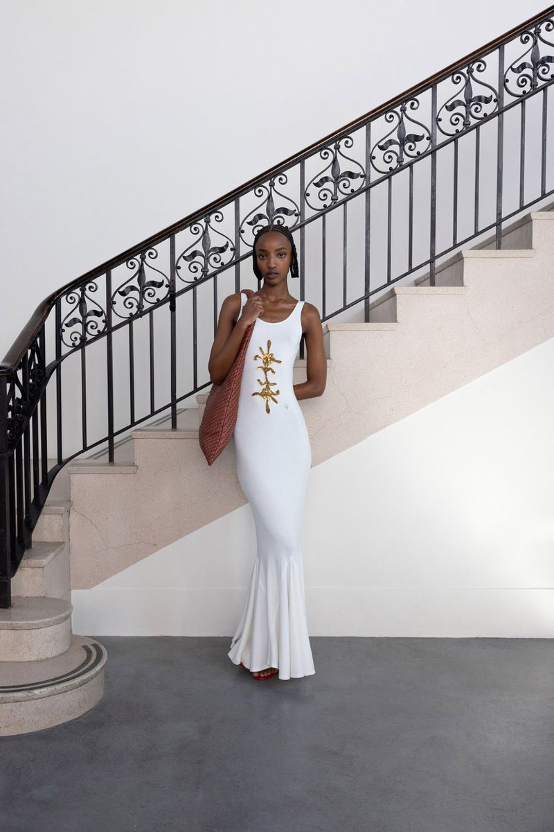 A woman in a white dress stands on a staircase with a patterned shoulder bag in an indoor setting. The photograph is well-lit.