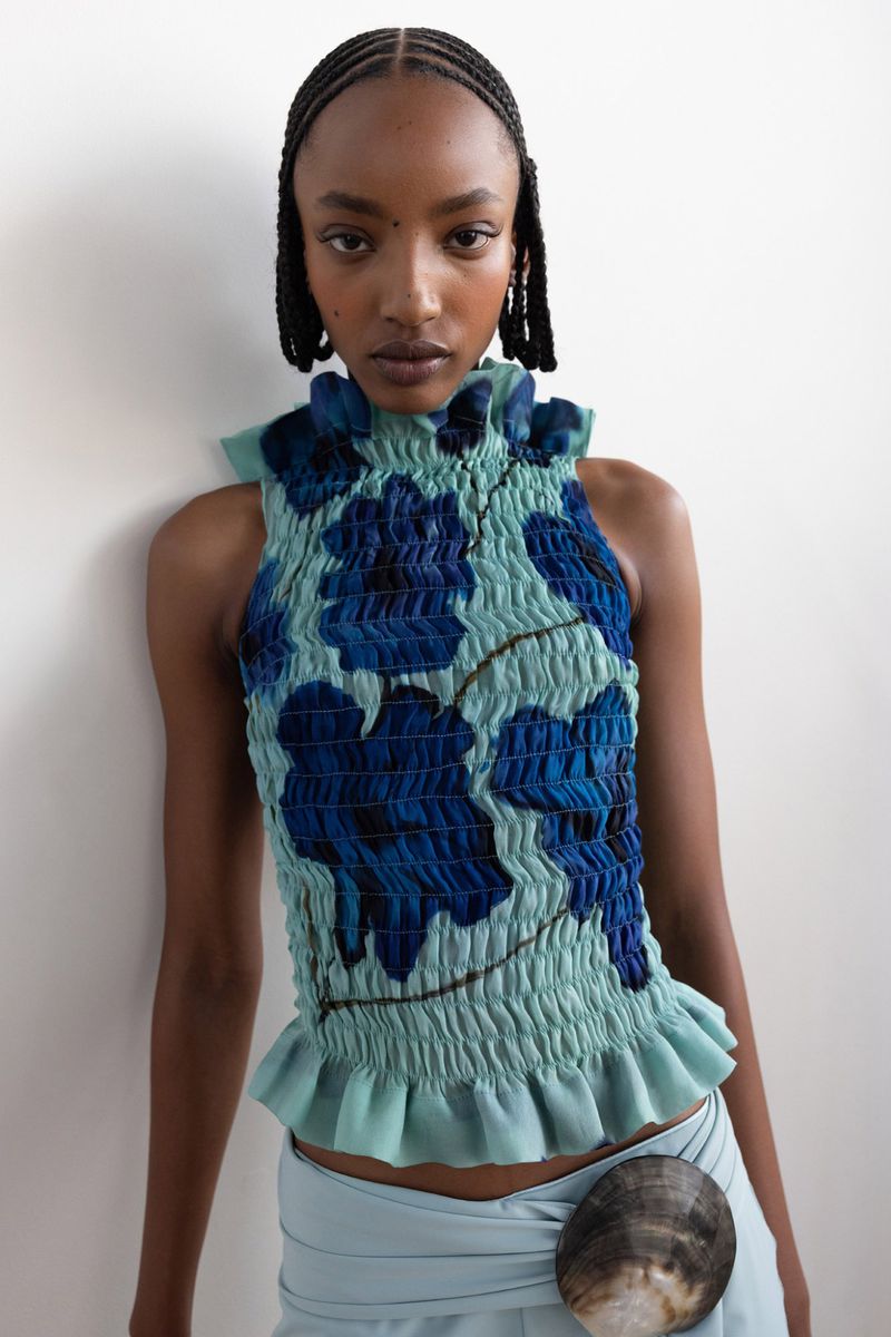 Fashion model wearing a light blue floral top and matching skirt with a shell accessory posing in a studio with a white wall backdrop.