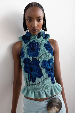 Fashion model wearing a light blue floral top and matching skirt with a shell accessory posing in a studio with a white wall backdrop.