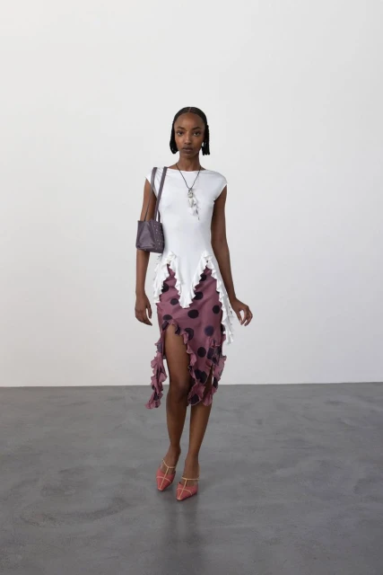 A slender Black woman poses in a white top and purple polka-dot skirt with red shoes, carrying a small purse in a minimalist studio setting.