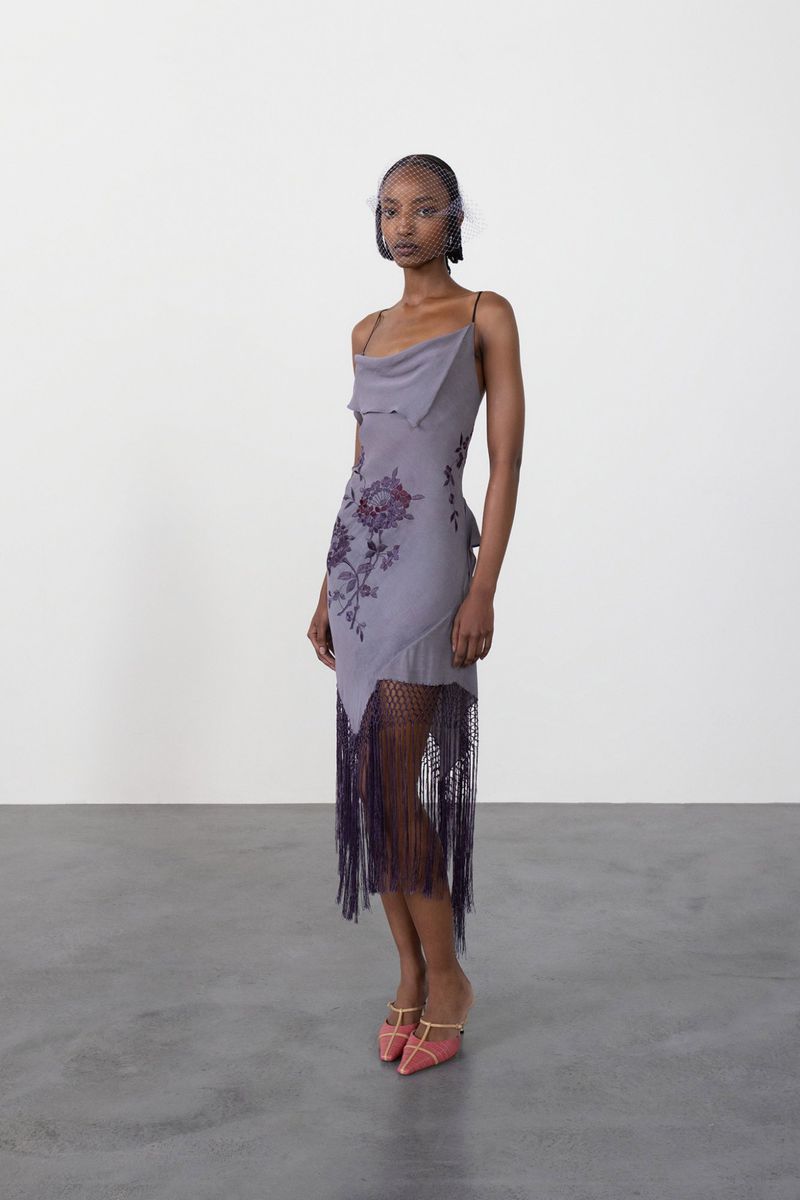 Elegant fashion shot of a woman in a gray dress with fringe and floral details, accessorized with a net headpiece and pink heels, indoors.
