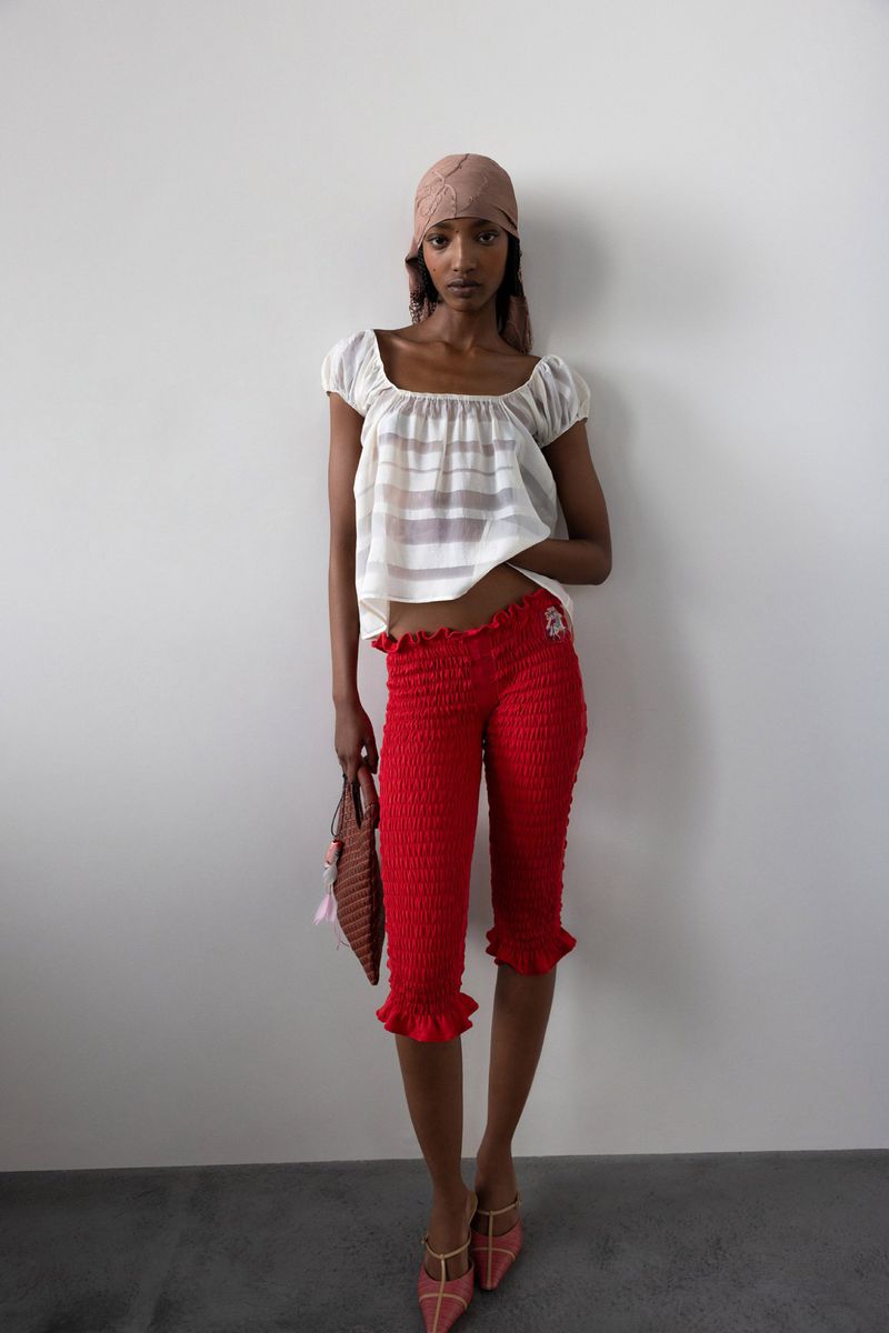 A dark-skinned young woman models a cream-colored peasant blouse, red ruffled capri pants, headscarf, and brown purse against a white wall.