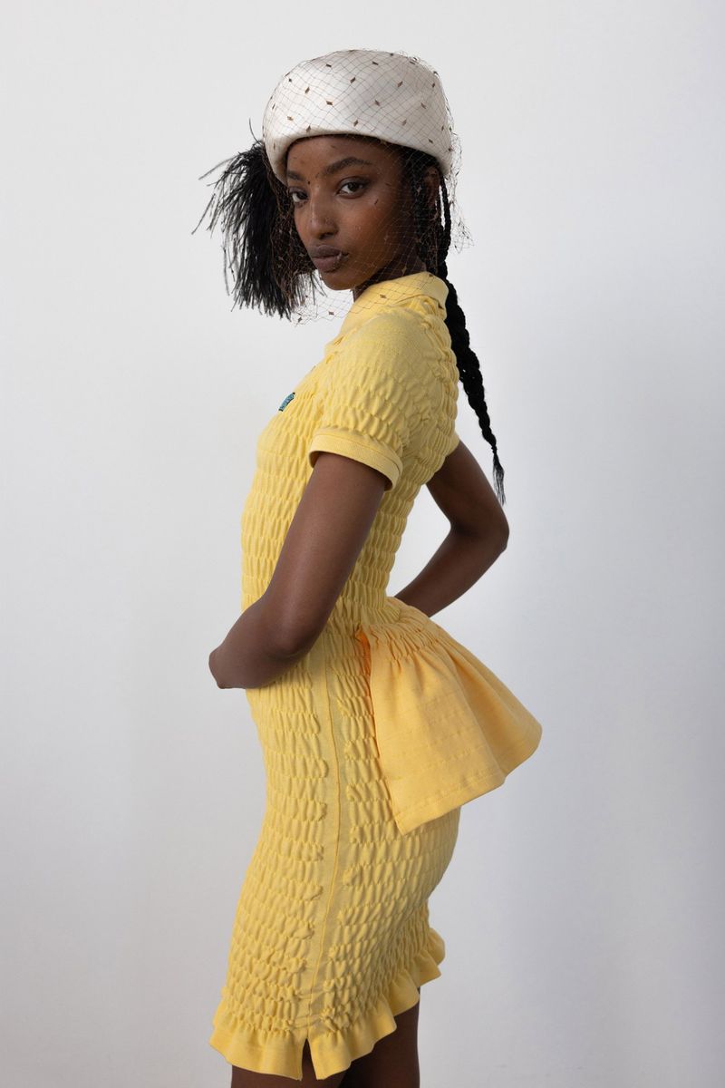 Fashionable woman in yellow dress and hat poses against a white backdrop in a studio setting; stylish, contemporary design.