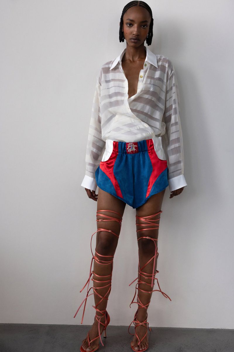 Woman in striped blouse and color-blocked shorts with wrap-around sandals strikes a pose against a neutral backdrop in a fashion forward ensemble.