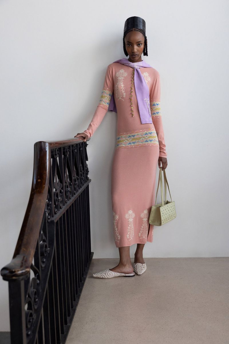 Woman in a peach-colored dress, lilac scarf, and black cap stands on a stairwell. She holds a purse. Fashion photography.