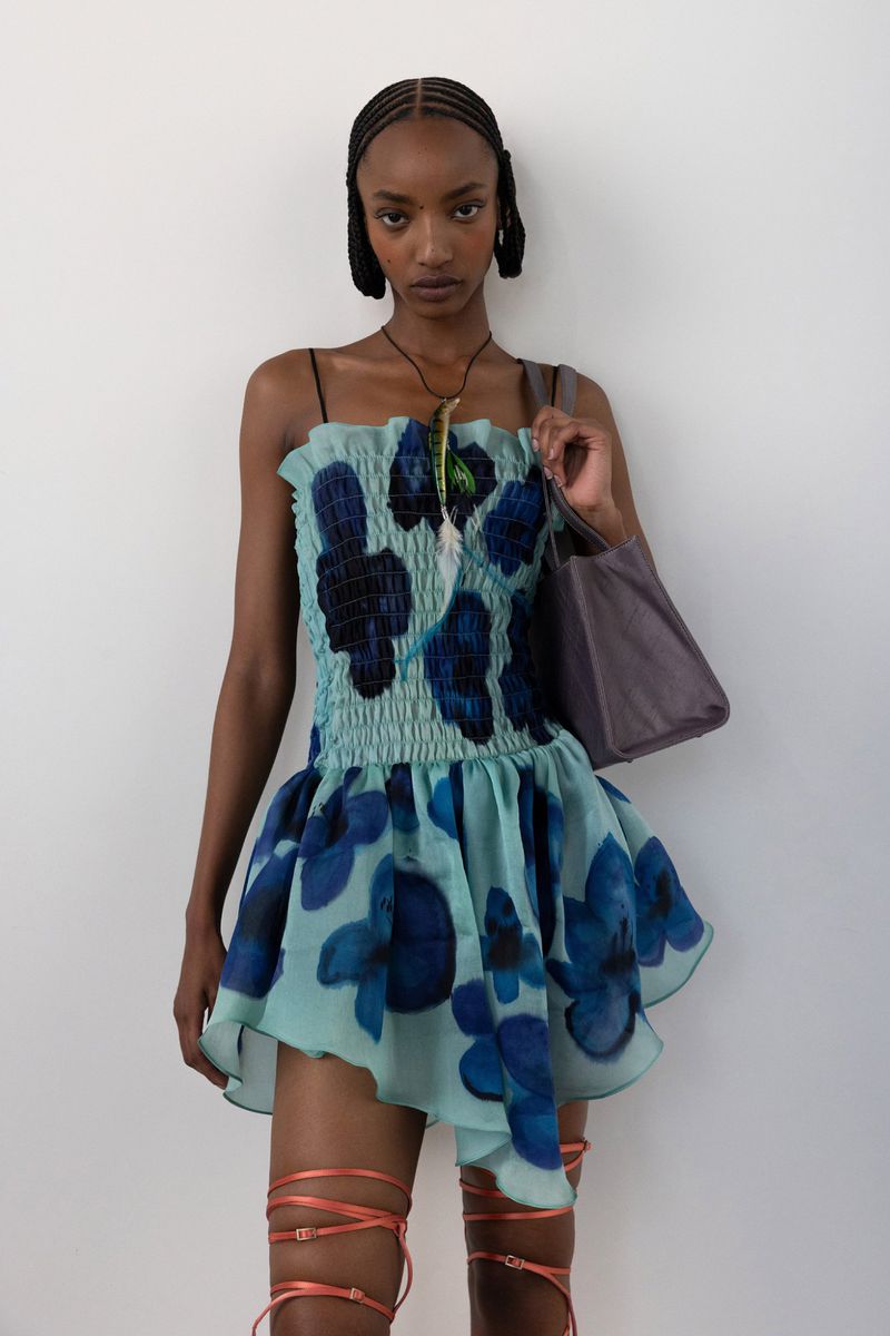 A woman poses in a blue floral mini dress against a white background, holding a tote bag, and wearing strappy sandals.