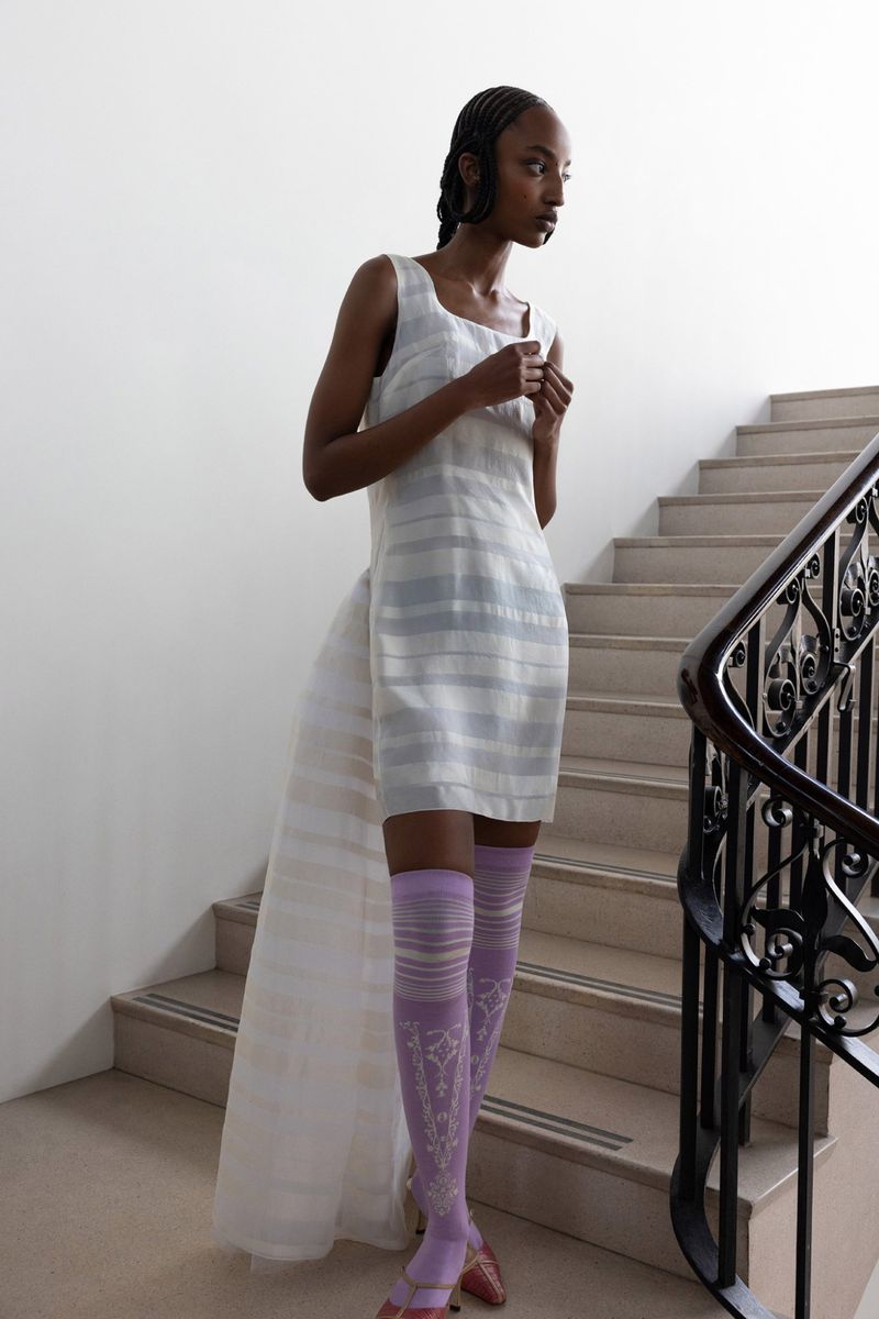 A slender woman poses on stairs wearing a striped mini dress with a train, lavender socks, and strappy sandals, looking right.