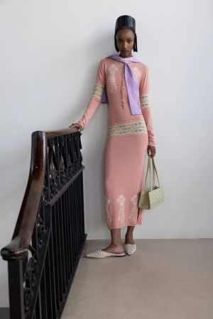 Model wearing a pink patterned dress and a lavender sweater over her shoulders standing beside a dark ornate staircase railing.