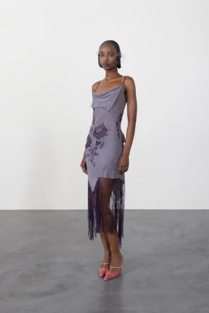 Black model in a cowl-neck lavender slip dress with floral embroidery and fringe hem standing against a grey backdrop.