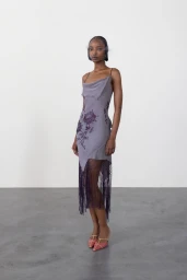 Black model in a cowl-neck lavender slip dress with floral embroidery and fringe hem standing against a grey backdrop.