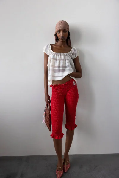Model wearing a white sheer top and red textured trousers holding a small handbag against a plain background