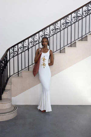 Model wearing a white dress with gold embellishment stands before a grand staircase for Conner Ives lookbook