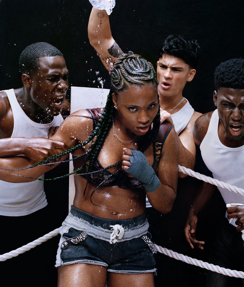 Female boxer surrounded by a cheering group with a water bottle being poured. She wears a sports bra, shorts, and boxing hand wraps.