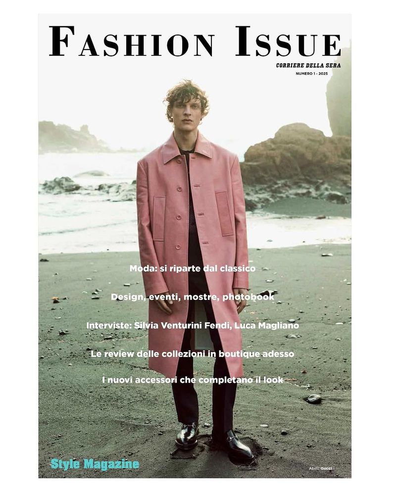 Magazine cover featuring a fair-skinned male model in a pink coat, standing on a beach with a rocky coastline backdrop.