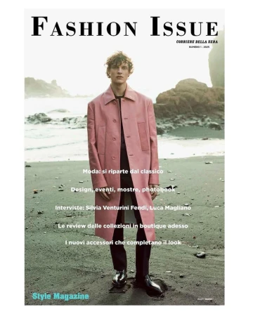 Magazine cover featuring a fair-skinned male model in a pink coat, standing on a beach with a rocky coastline backdrop.