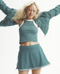 Blonde woman wearing a green quilted jacket, matching tank top, and skirt against a white studio background.