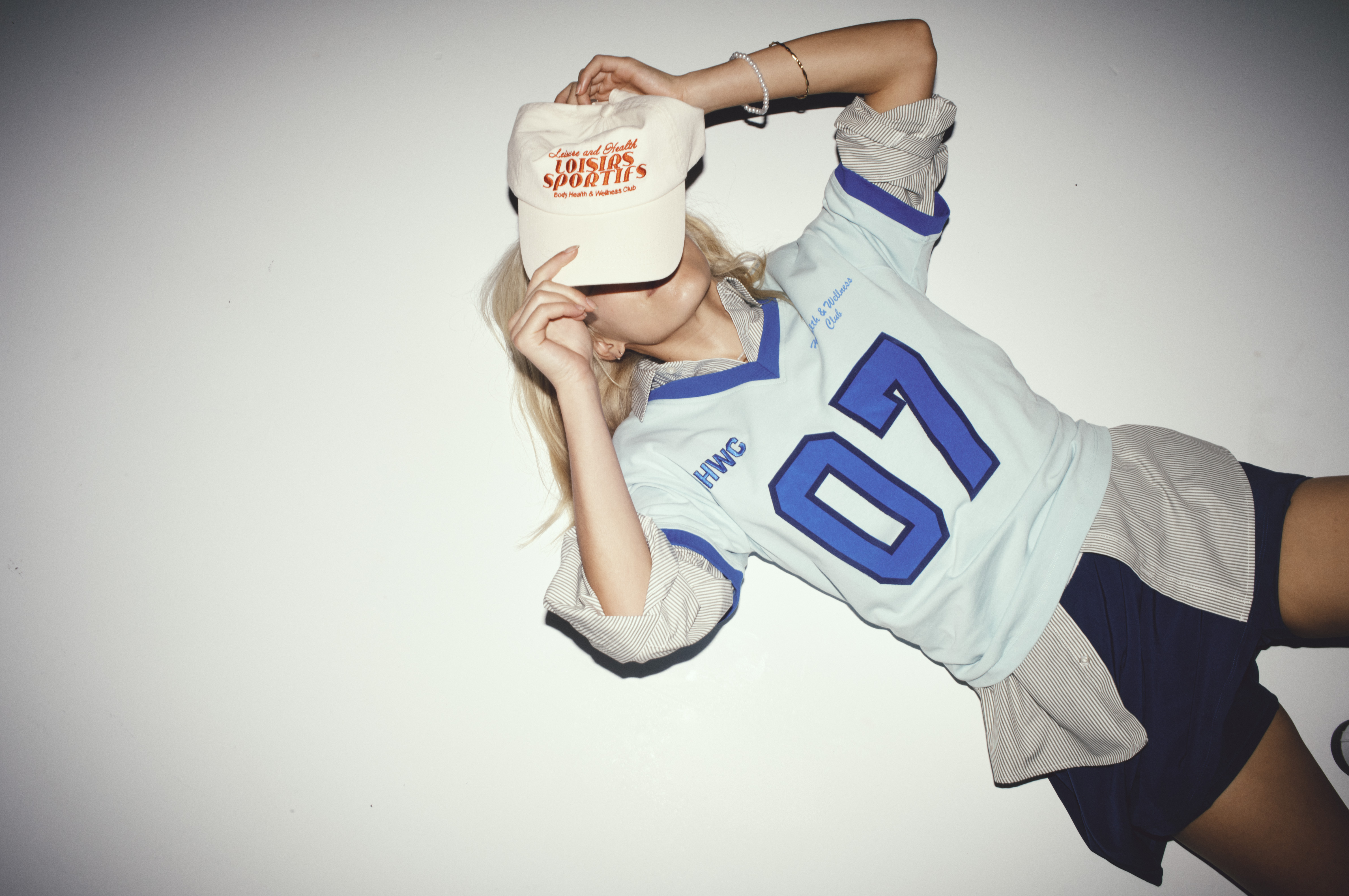 Young woman in athletic wear poses against a white wall, covering her face with a baseball cap with red text.