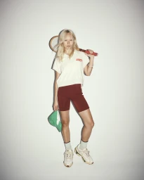 Model with blonde hair holding a tennis racket over her shoulders against a plain grey studio background.