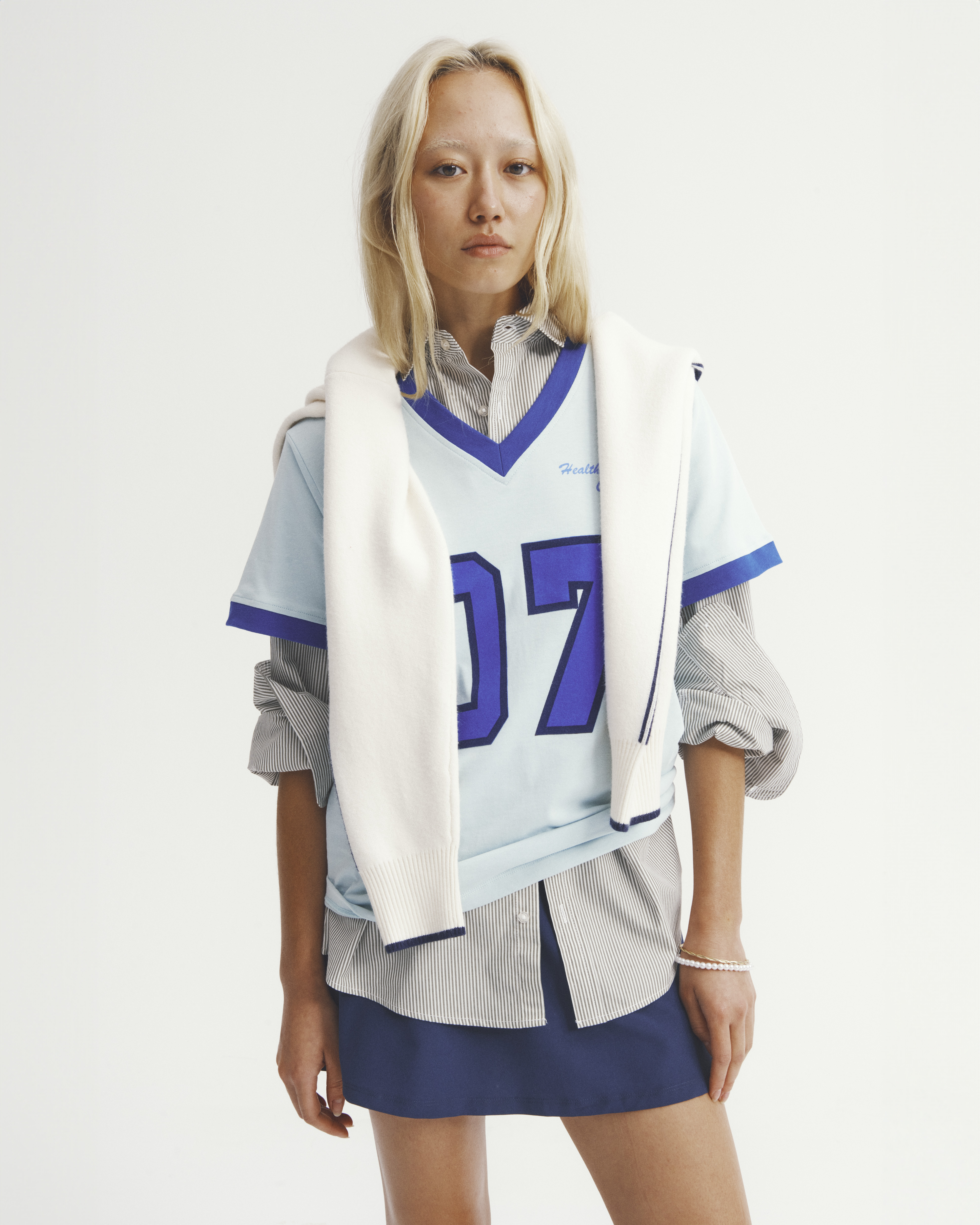 Young woman in layered fashion wearing a pale blue jersey with a white sweater draped over the shoulders against a white backdrop.