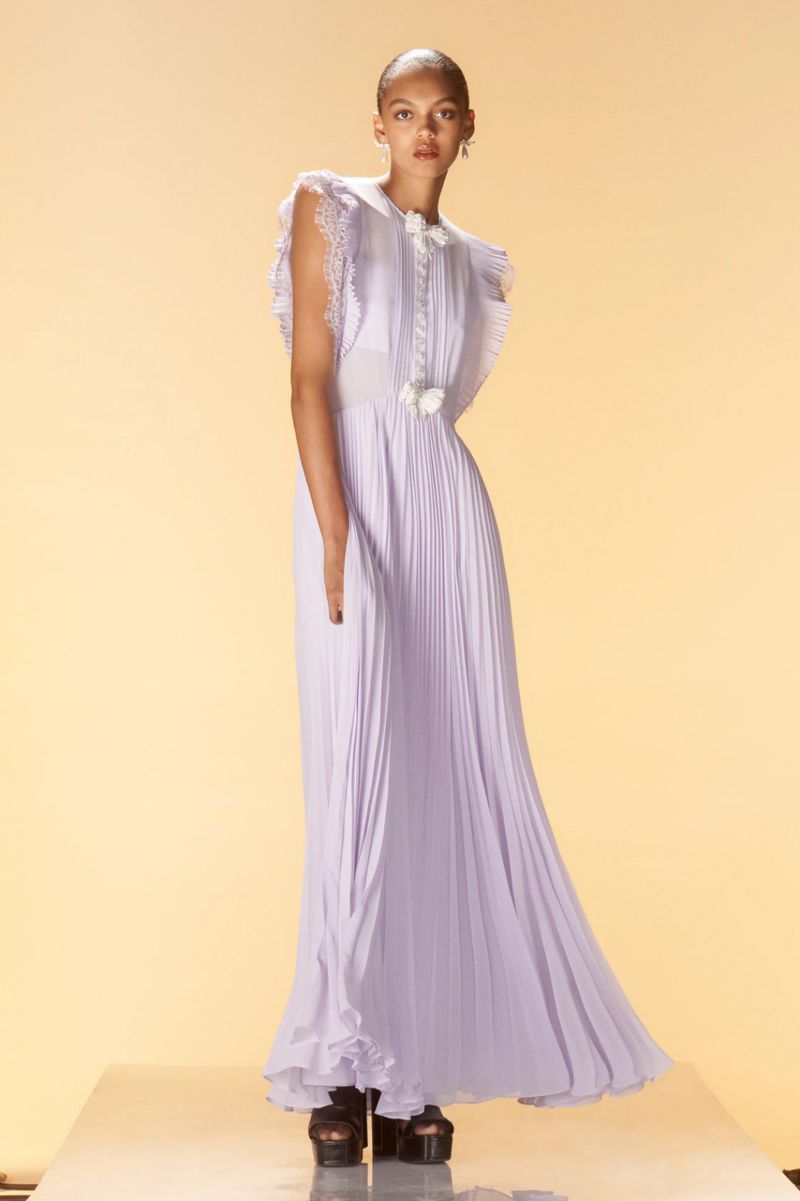 A lone fashion model poses in a floor-length periwinkle gown with pleats, lace details, and cap sleeves against a muted yellow backdrop.