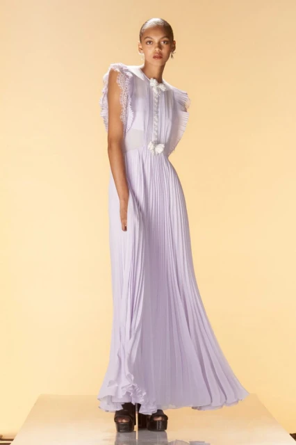 A lone fashion model poses in a floor-length periwinkle gown with pleats, lace details, and cap sleeves against a muted yellow backdrop.