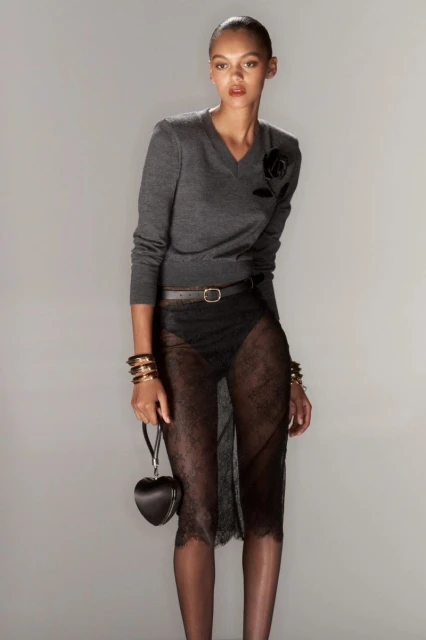 Fashion shot of a woman with a gray sweater, black belt, sheer lace skirt, dark bracelets, and a heart-shaped purse against a gray background.