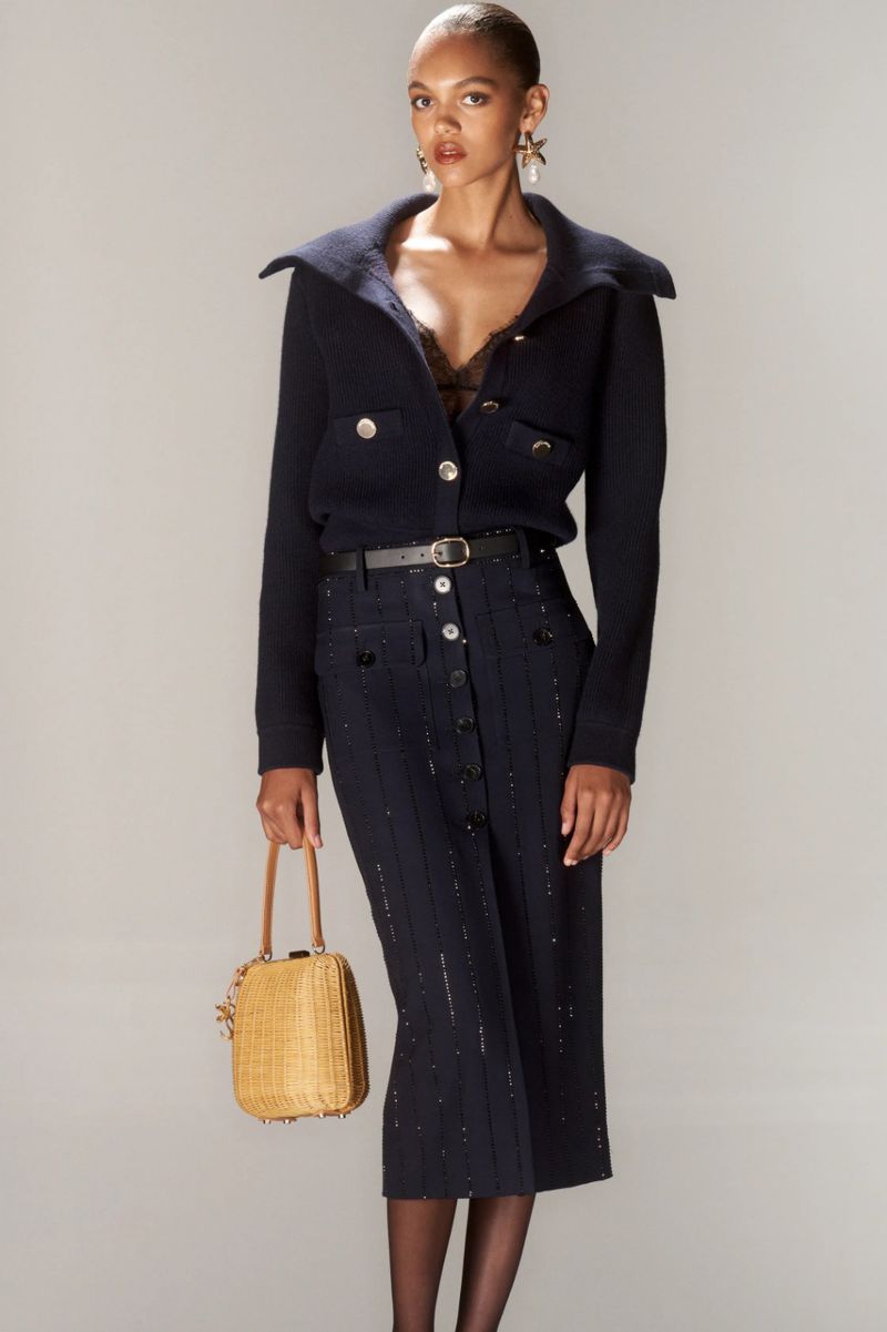 Elegant Black woman in stylish navy knit jacket, button-front pencil skirt, and woven purse poses in a neutral studio setting.