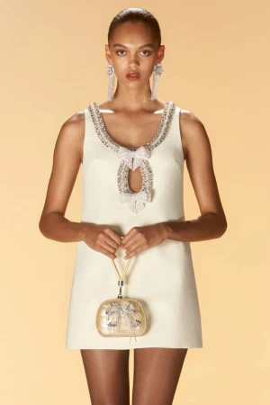 Fashion shot of a dark-skinned model wearing a cream-colored mini dress with bejeweled trim, holding a handbag, and posing indoors.